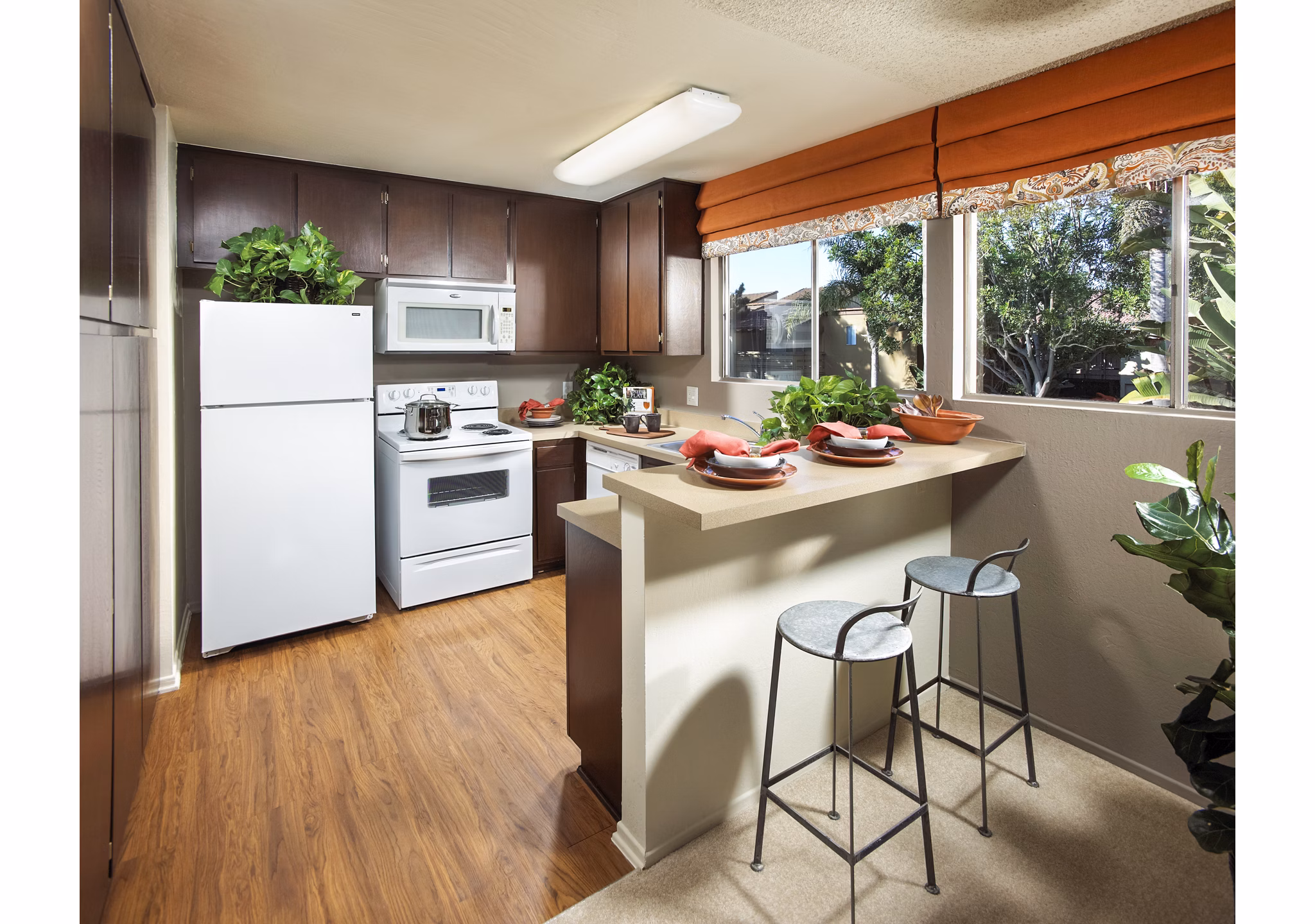 Modern kitchen with dark wood cabinets and a small bar with stools, featuring white appliances and decorative plants