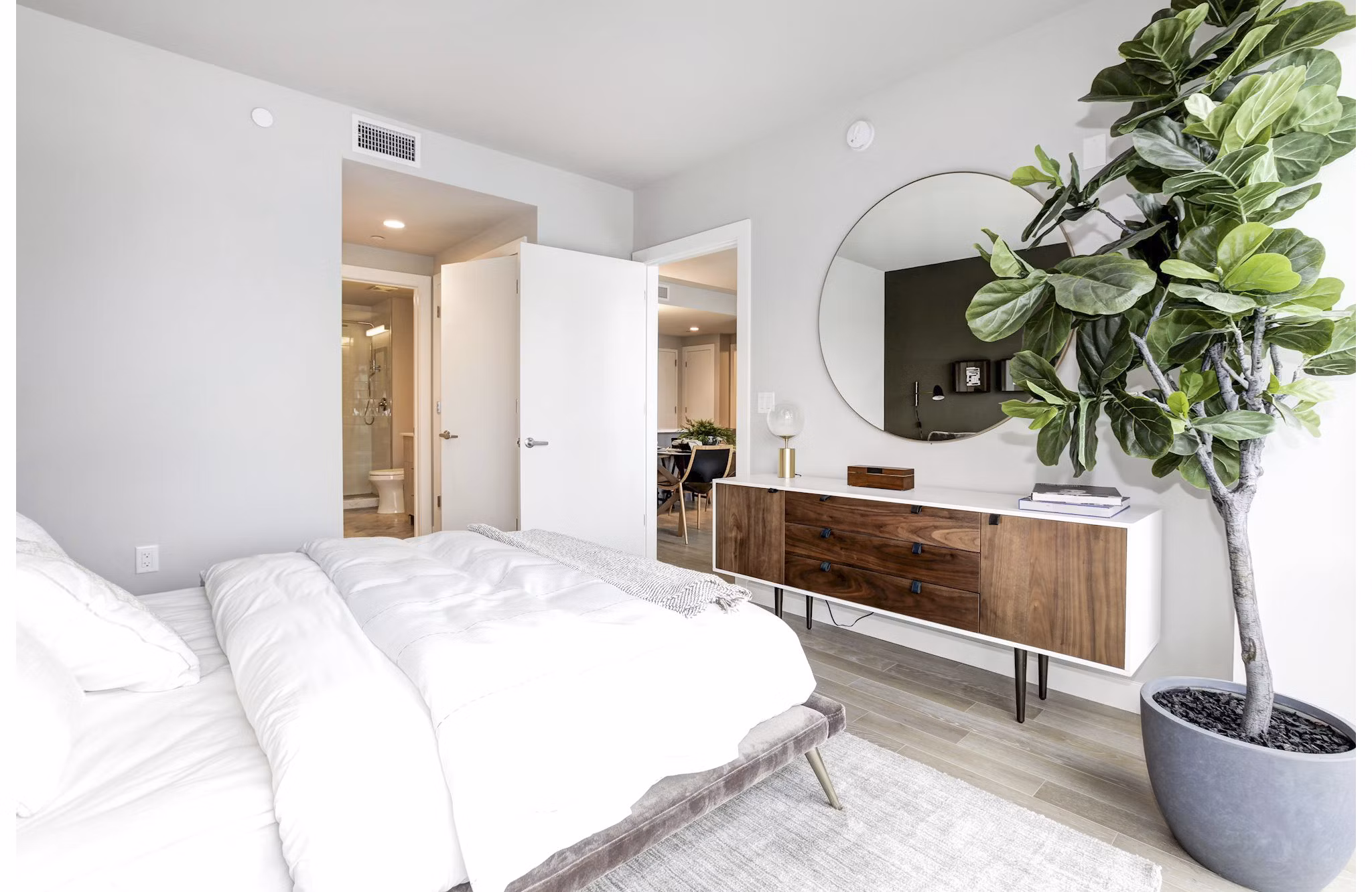Bright bedroom with large plant, round mirror, and wooden dresser at Harbor 1500 Apartments