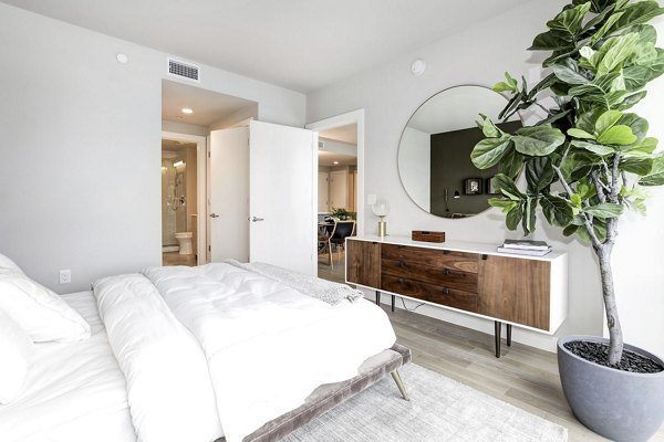 Bright bedroom with large plant, round mirror, and wooden dresser at Harbor 1500 Apartments