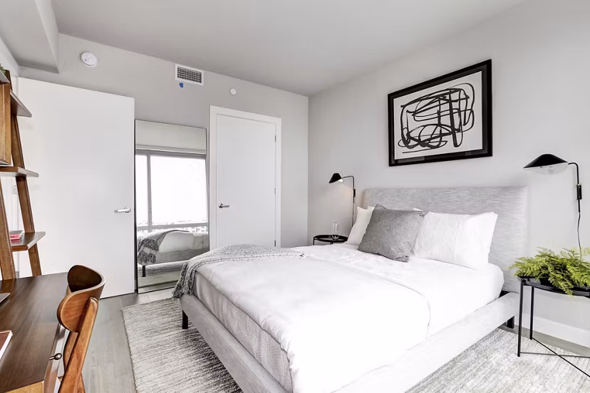 Minimalist bedroom with white bed, abstract art, and desk at Harbor 1500 Apartments in Weehawken, offering serene luxury living spaces