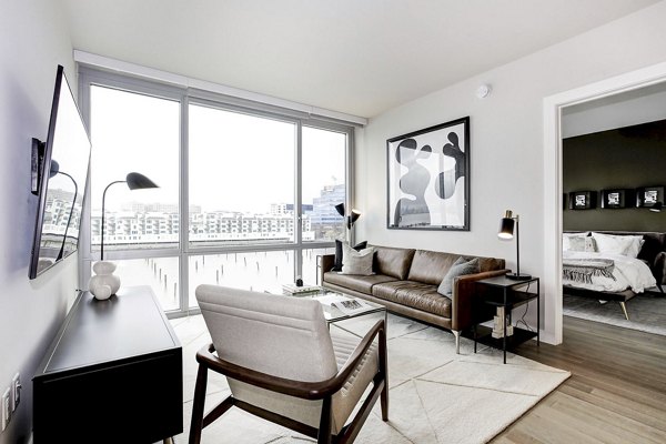 Bright living room with large windows, leather sofa, and artwork in Harbor 1500 Apartments, luxury living by Greystar