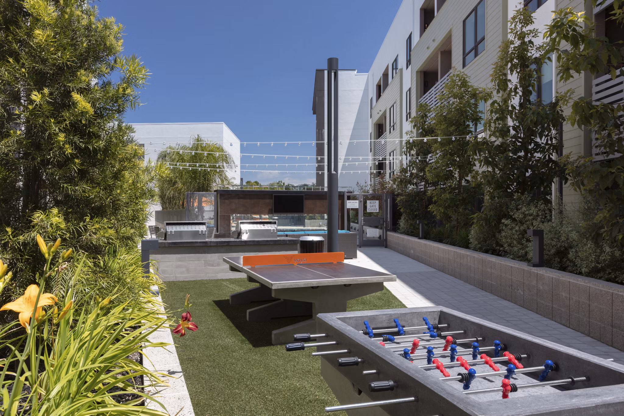 Outdoor recreational area with ping pong and foosball tables surrounded by greenery and seating at luxury apartments