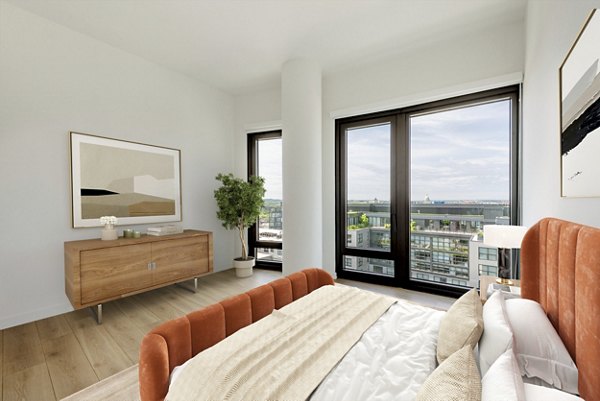 Modern bedroom featuring a bed, wooden dresser, plant, and large windows with city skyline views