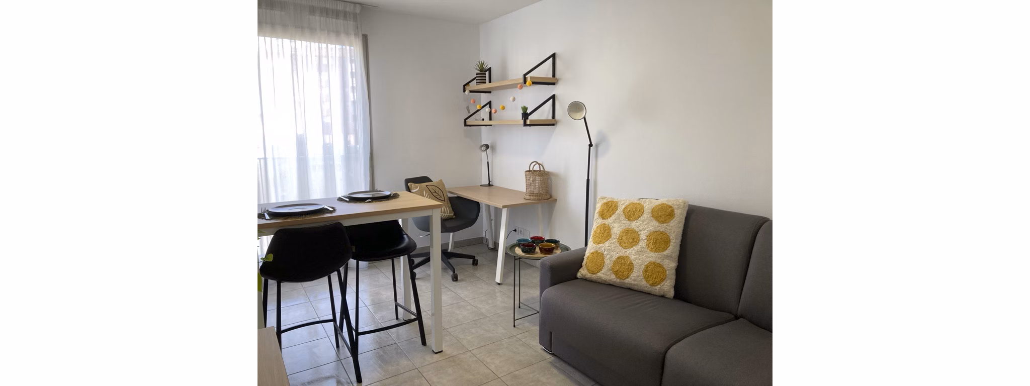 Modern living room with gray sofa, yellow accent pillow, coffee table, and decorative shelving, featuring a large window letting in natural light.