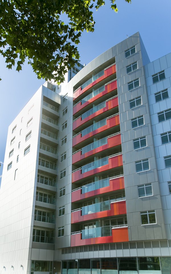 Modern apartment building exterior with a red and gray facade, featuring large windows and a contemporary architectural design surrounded by trees.