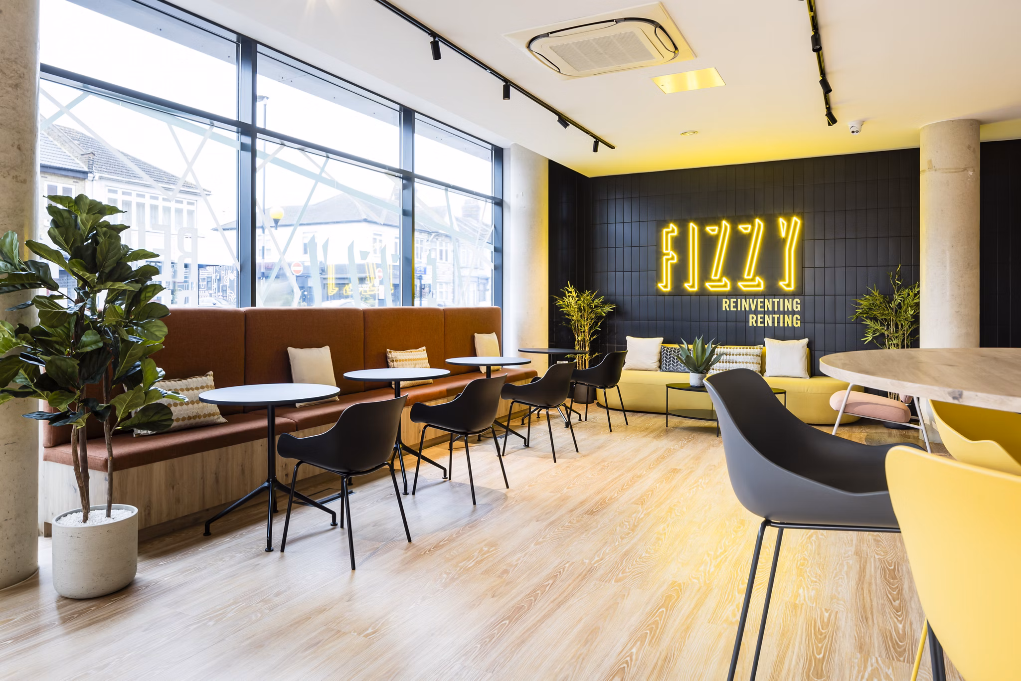 Bright communal lounge at Fizzy Walthamstow featuring a long table, vibrant decor, and large windows with natural light.
