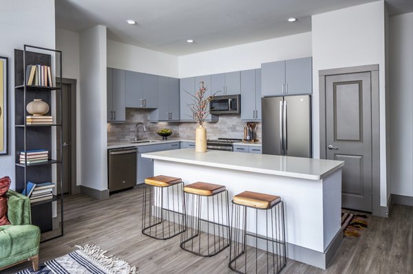 Modern kitchen showcasing gray cabinets, a white island with three bar stools, and stainless steel appliances in luxury apartments
