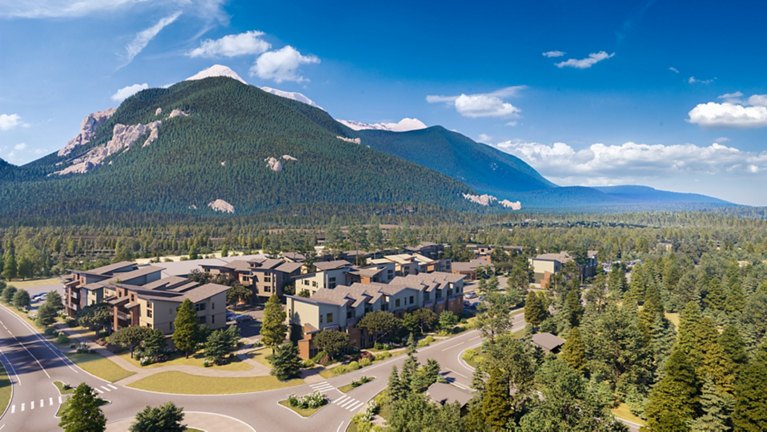 Aerial view of a residential community with multi-story buildings nestled in a lush forest at the base of a mountain range under a clear blue sky.