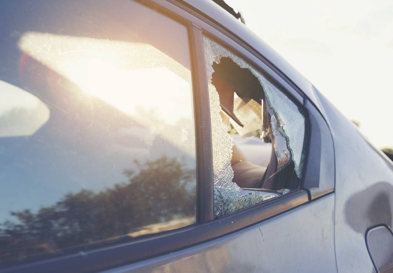 Close-up of a car's shattered side window with glass fragments around the frame, under bright sunlight.