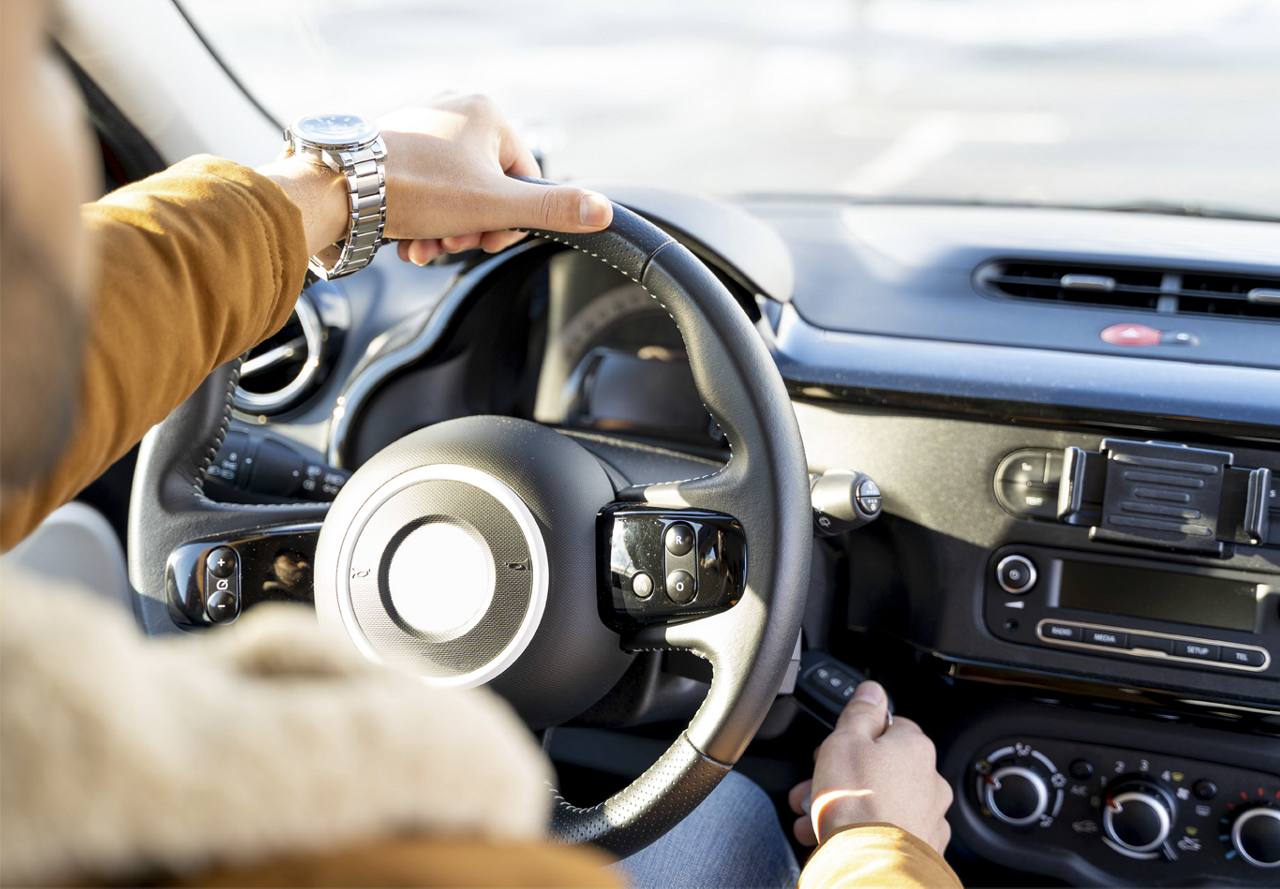 Person's hands on a car steering wheel with a wristwatch visible, and the car's dashboard and ignition in the background.