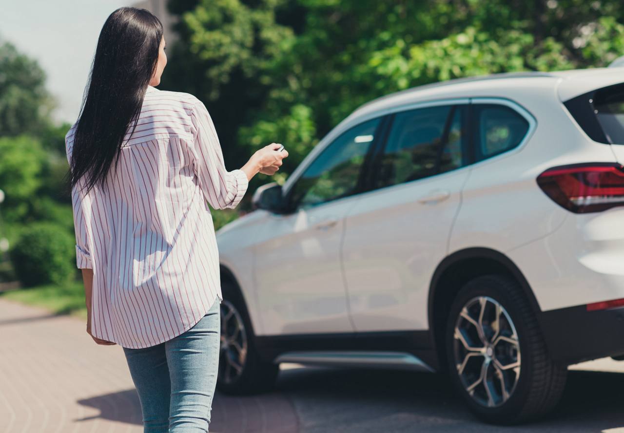 A woman from behind walking towards a parked car while holding car keys, ready to unlock it.