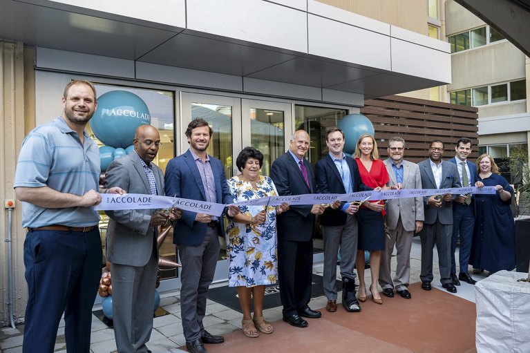 A group of individuals partaking in a ribbon-cutting ceremony with a banner reading 'ACCOLADE', signifying a celebratory event or opening.