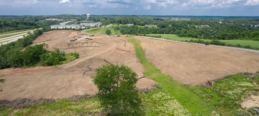 Aerial view of a large, cleared construction site with earth-moving equipment, adjacent to a highway, set against a backdrop of greenery and a cloudy sky.