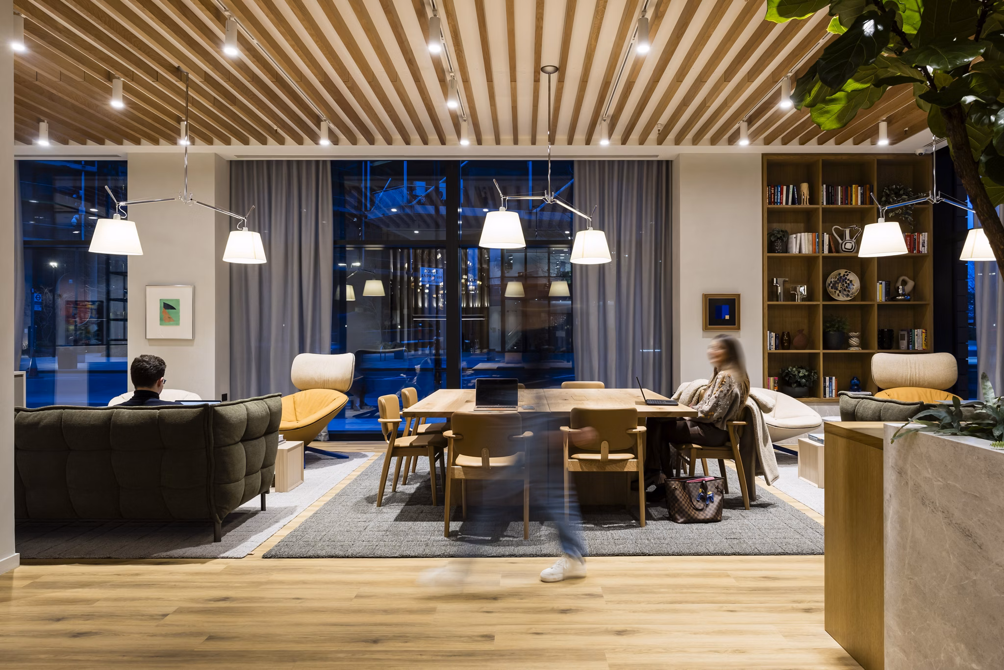 Modern and stylish common area at Bloom East Apartments in London, featuring comfortable seating, a wooden ceiling, bookshelves, and warm lighting.