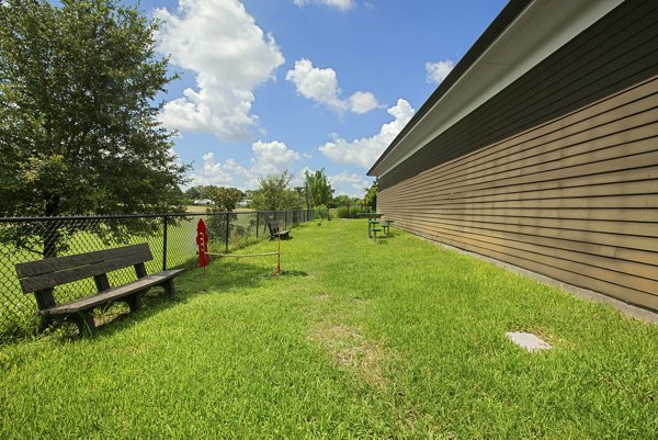 Grassy area with wooden bench and trees near luxury apartments under a partly cloudy sky