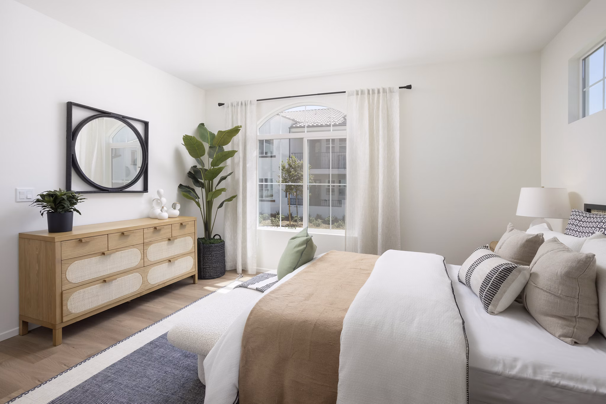 Modern bedroom featuring a bed, dresser with mirror, potted plant, and natural light from window curtains