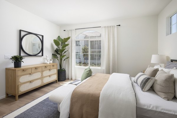Modern bedroom featuring a bed, dresser with mirror, potted plant, and natural light from window curtains