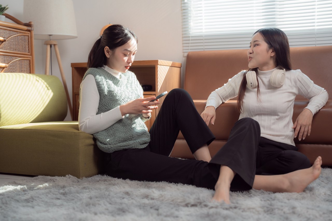 Two young adult asian women are sitting on a carpet in the living room, one is using a smartphone and the other is listening, wearing headphones around her neck