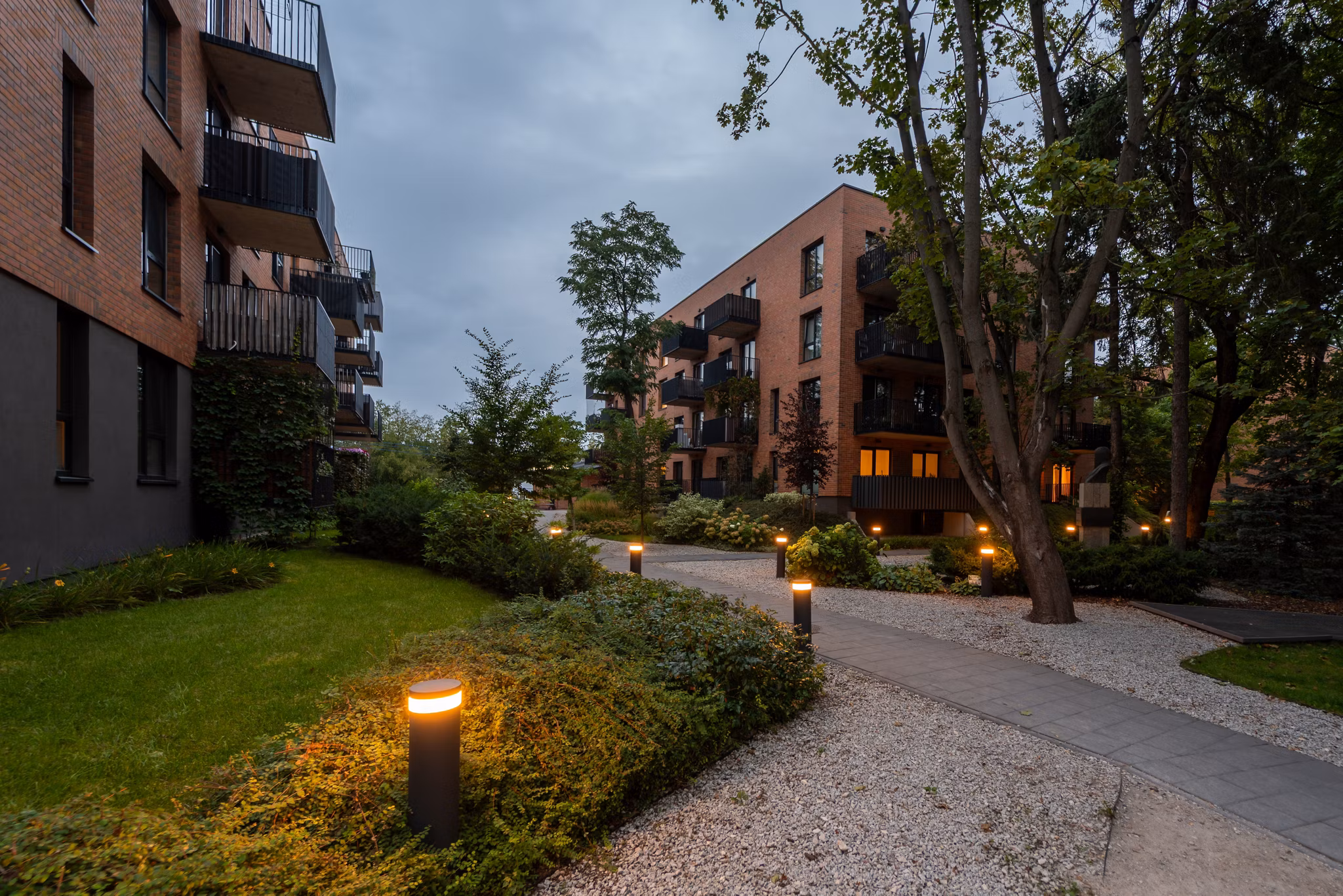 Illuminated pathway in landscaped courtyard of modern residential complex at dusk. Bollard lights create warm ambiance among lawns, shrubs, trees, near apartment buildings. A peaceful community space.