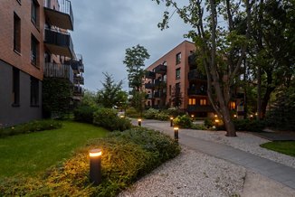 Illuminated pathway in landscaped courtyard of modern residential complex at dusk. Bollard lights create warm ambiance among lawns, shrubs, trees, near apartment buildings. A peaceful community space.