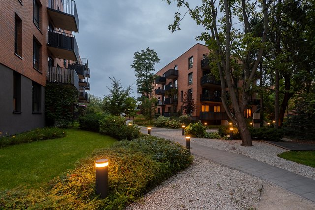 Illuminated pathway in landscaped courtyard of modern residential complex at dusk. Bollard lights create warm ambiance among lawns, shrubs, trees, near apartment buildings. A peaceful community space.