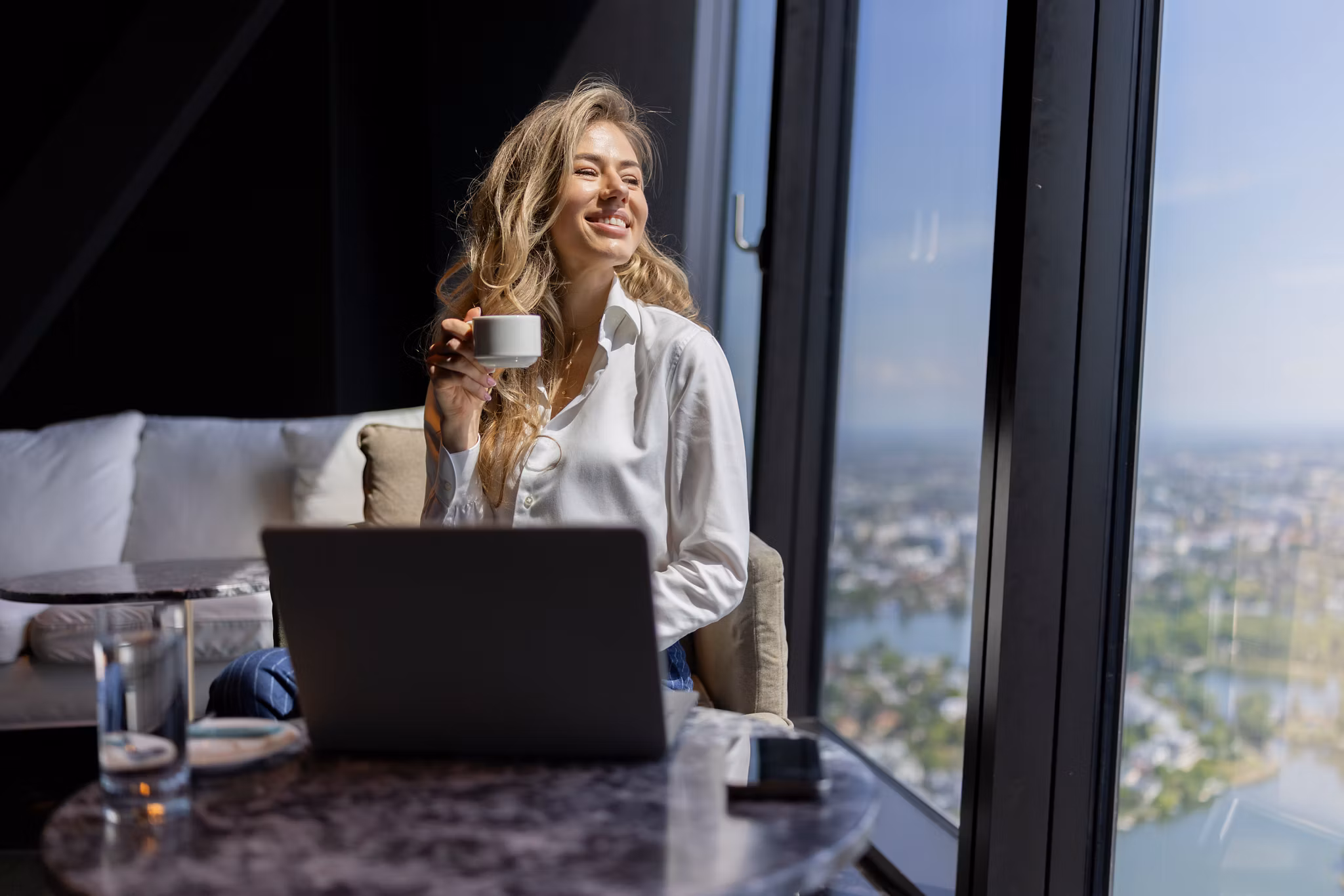 A joyful woman happily sipping coffee while working on her laptop in a stylish, modern highrise apartment, showcasing a breathtaking city view that inspires creativity and comfort