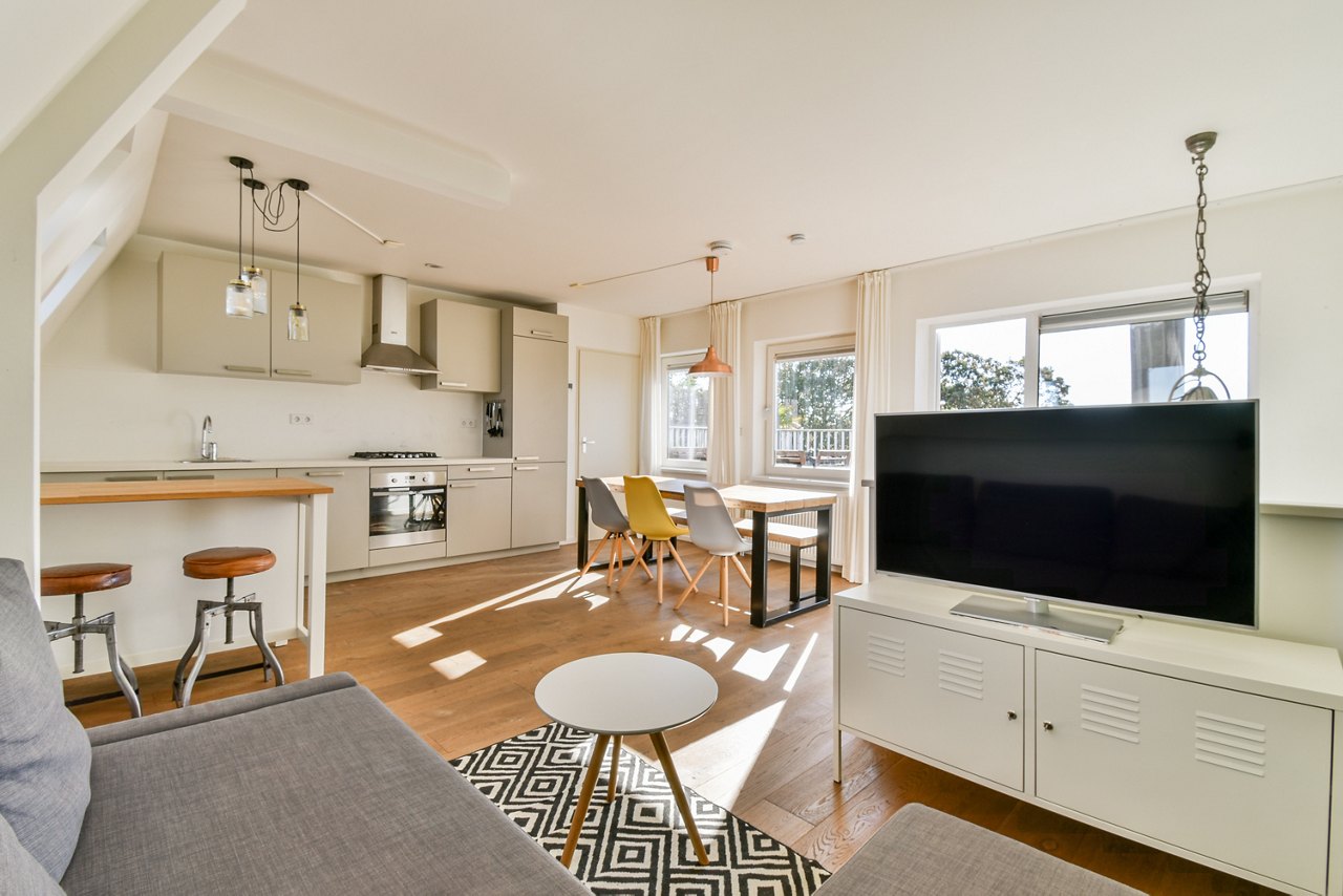 A stylish modern living room and kitchen area featuring minimalist decor, wooden flooring, and large windows for natural light.