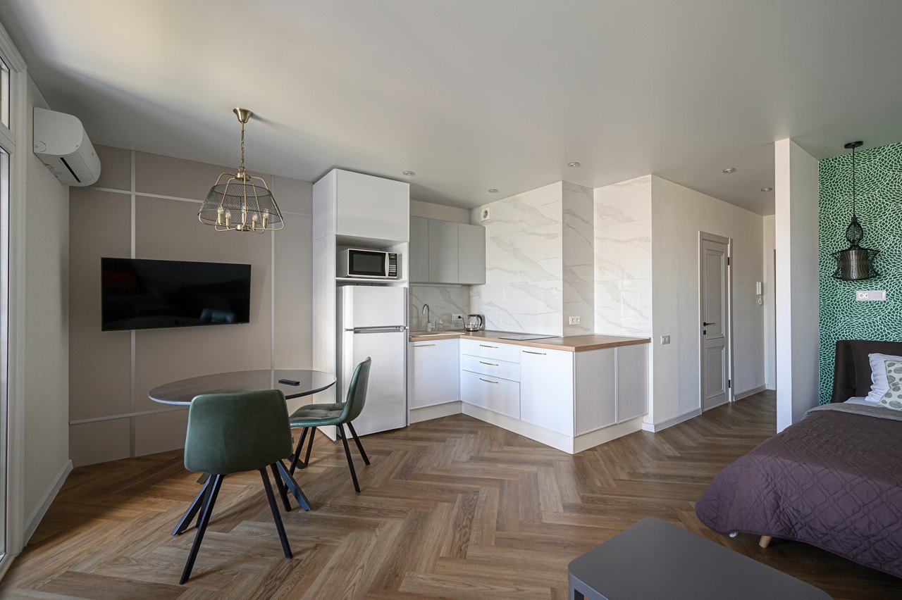 modern studio apartment featuring a small kitchen, dining area with green chairs, and a bed in a partitioned space. The interior design is clean and functional