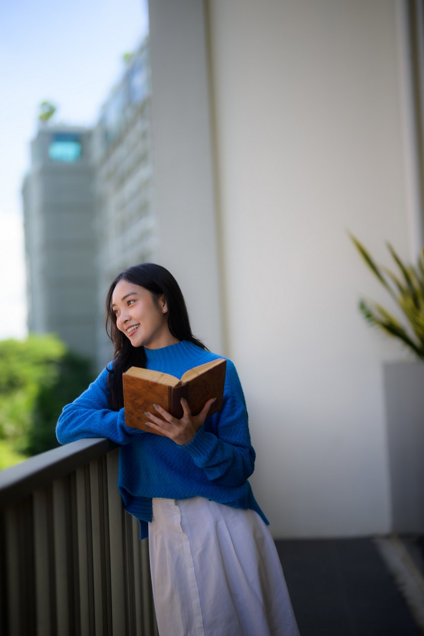 Young woman reading a book on a modern balcony, smiling and looking away, enjoying a moment of urban relaxation and contemplation