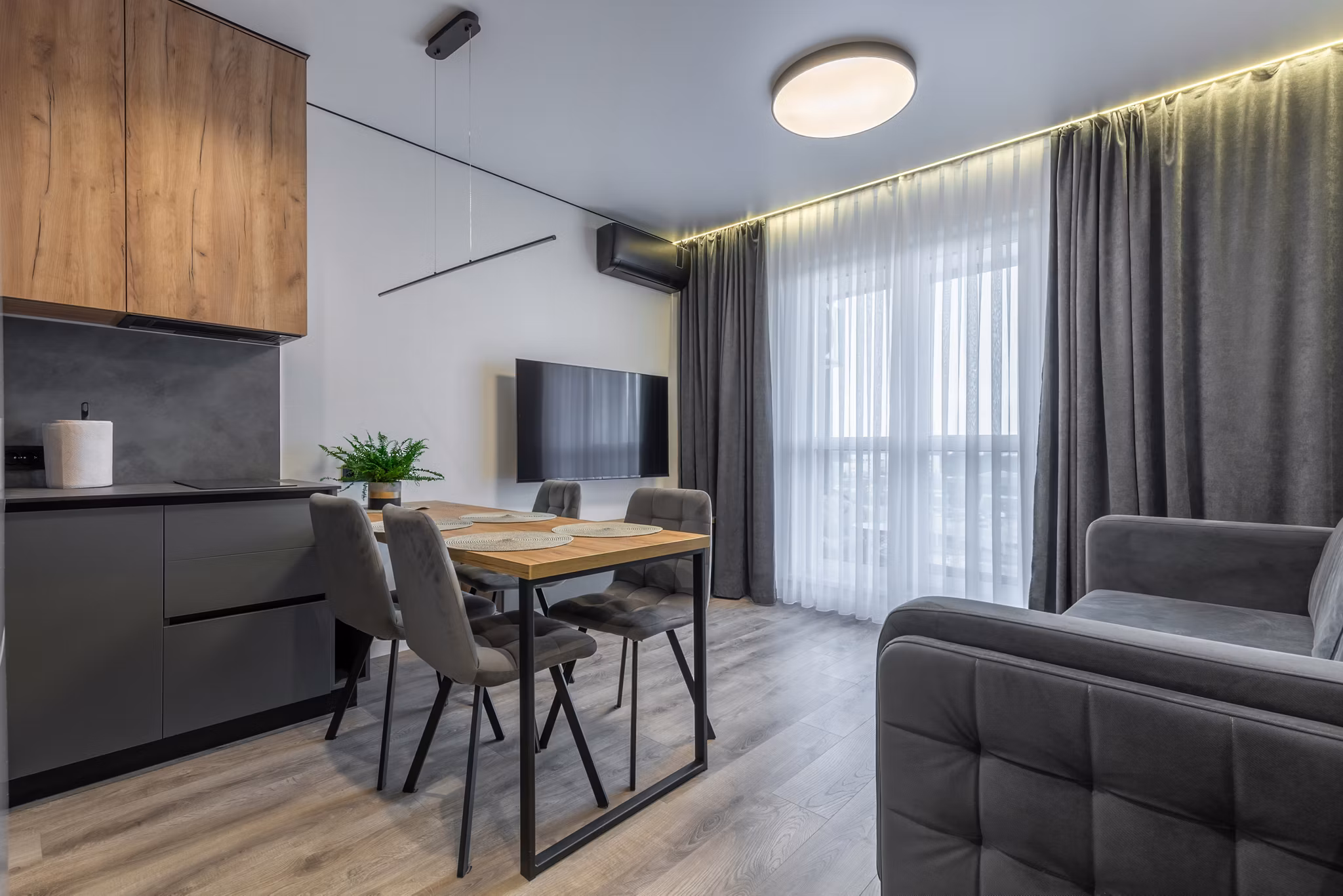 Contemporary open-plan space featuring a gray sofa, wooden dining table with upholstered chairs, sleek kitchen cabinets, and floor-to-ceiling window with sheer and blackout curtains.