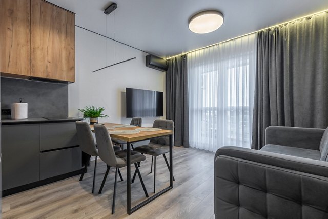 Contemporary open-plan space featuring a gray sofa, wooden dining table with upholstered chairs, sleek kitchen cabinets, and floor-to-ceiling window with sheer and blackout curtains.