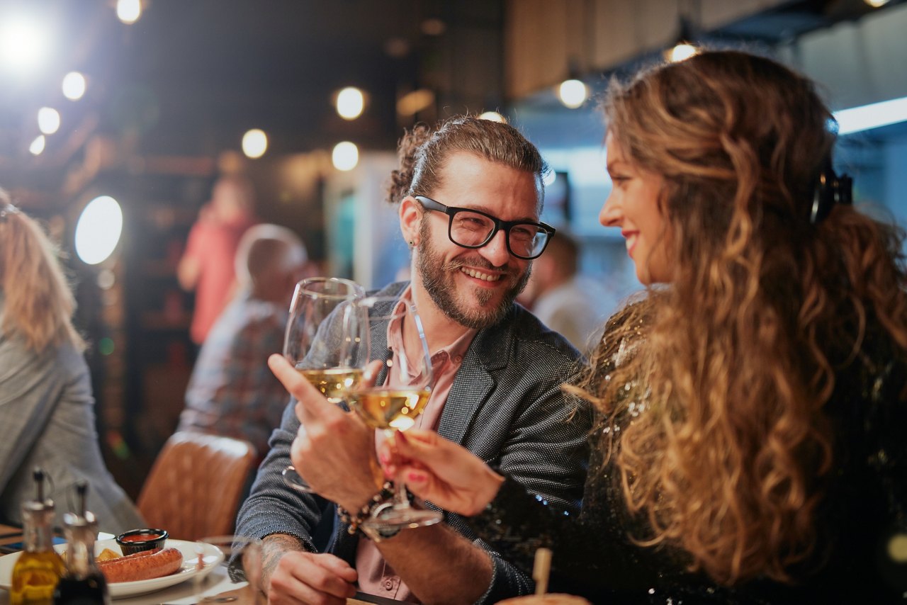 Cute caucasian couple sitting in restaurant and cheering with wine. In background are their friends.