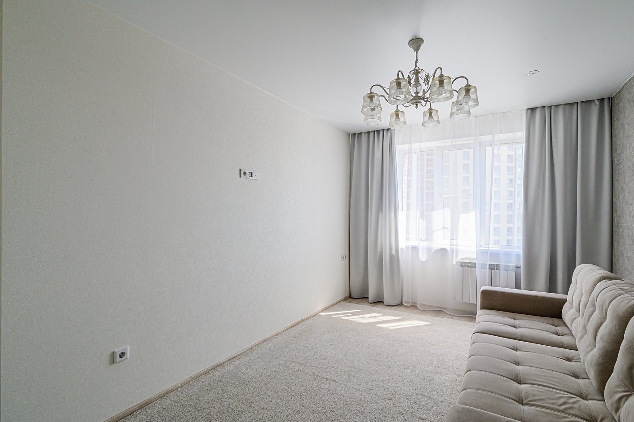 Cozy living room with light gray walls, soft carpet, and a comfortable beige sofa. Sunlight streams through the window with sheer and heavy curtains. A decorative chandelier hangs from the ceiling