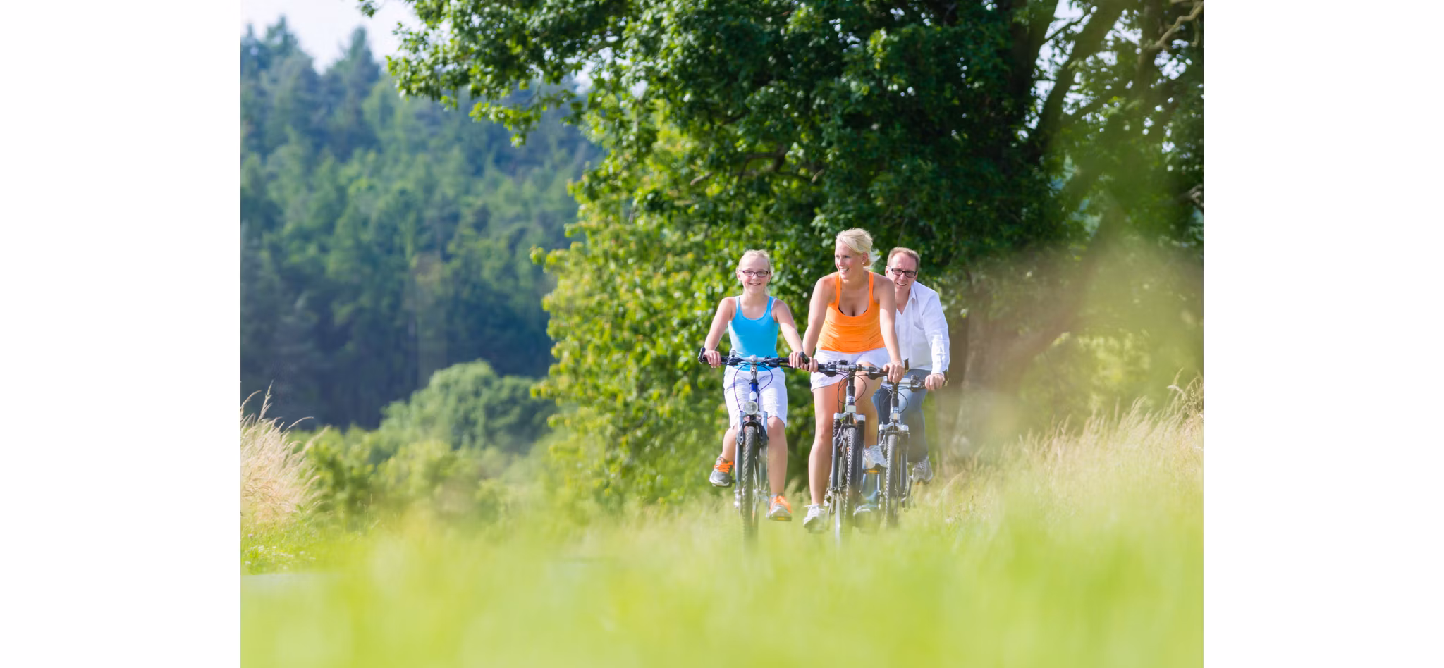 Three people cycling together on a grassy trail with lush green trees in the background on a sunny day.