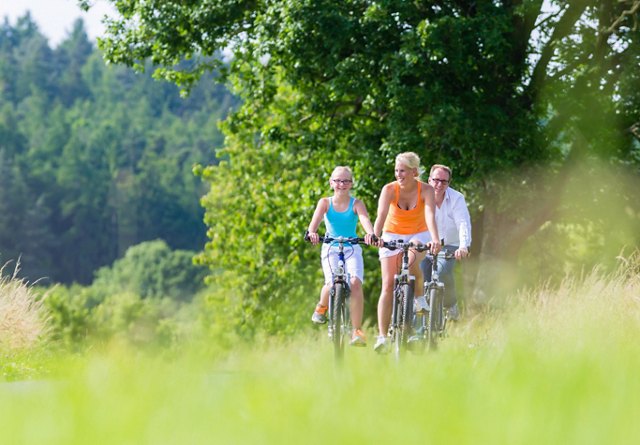 Three people cycling together on a grassy trail with lush green trees in the background on a sunny day.