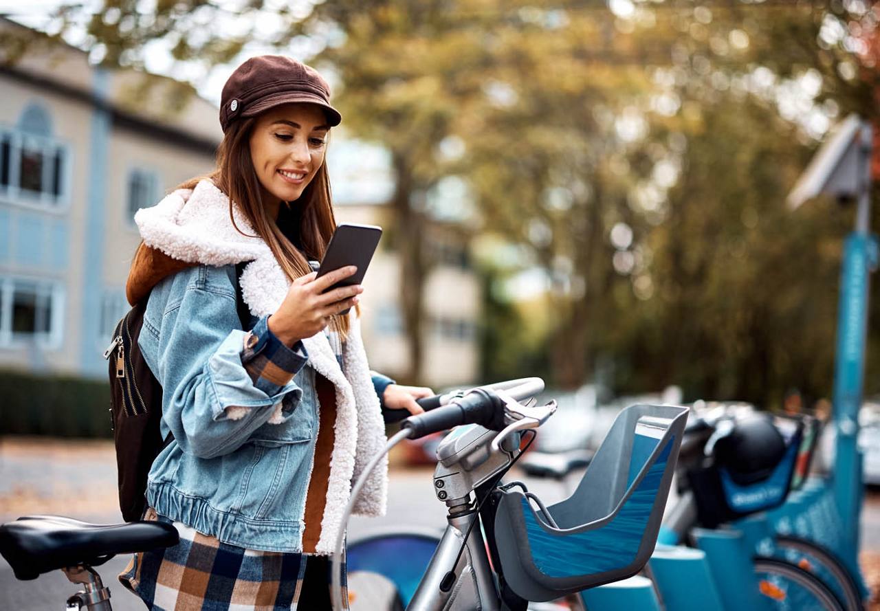 Smiling woman in a denim jacket and cap using a smartphone while standing by a rental bicycle station.