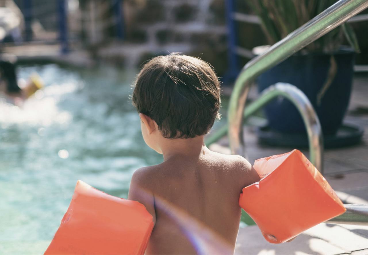 A young child with brown hair and orange arm floaties stands by a pool, looking at other swimmers.