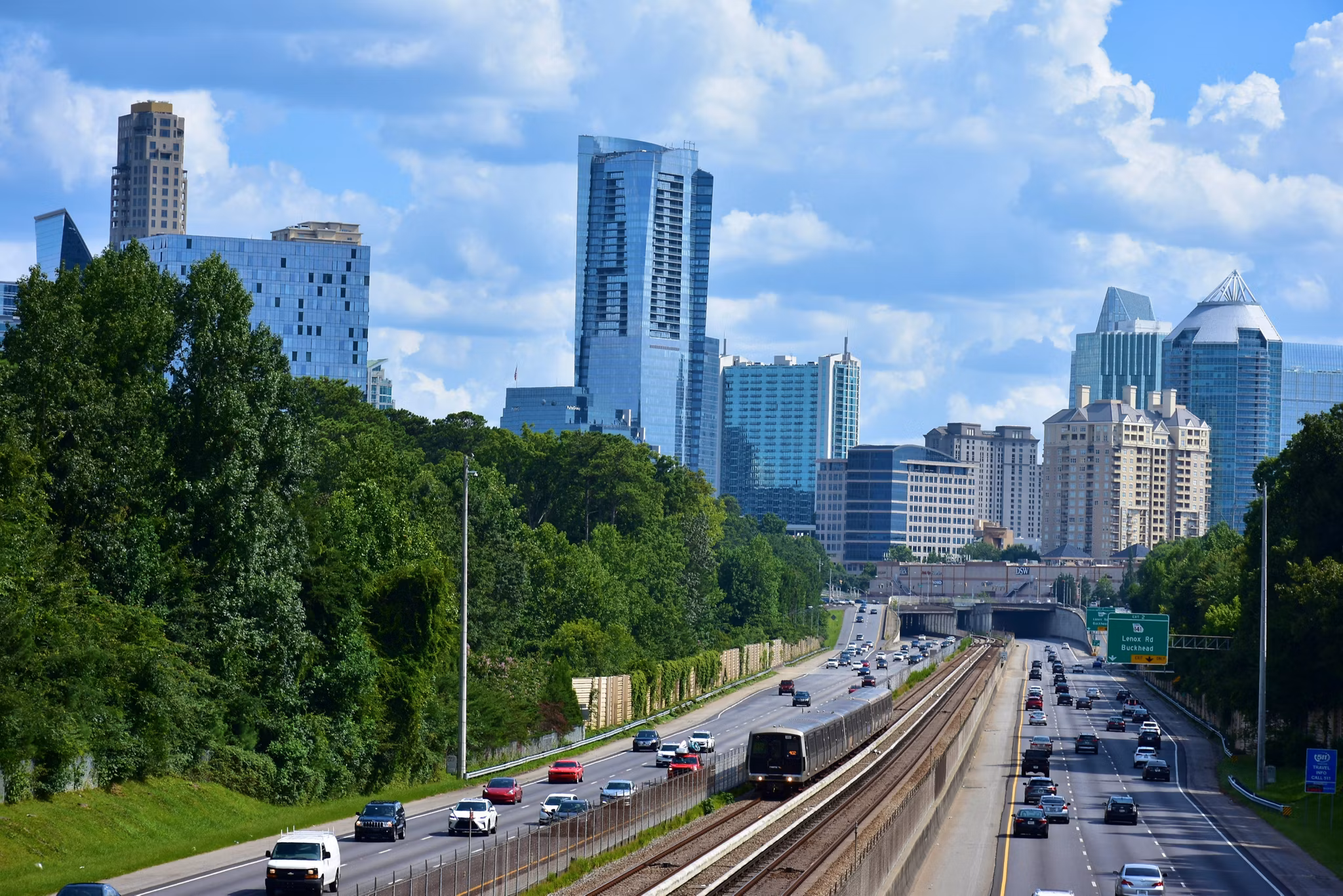 View of the Buckhead district skyline in Atlanta, Georgia, showing a MARTA heavy-rail train on the Red/Gold line corridor paralleling GA-400, with the Lenox Road and Buckhead highway exit signage visible. Illustrates transportation and commuting options for Atlanta residents.