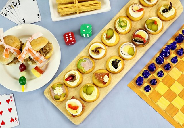 Assorted appetizers on a wooden board with sandwiches, colorful dice, playing cards, and a chessboard on a blue tablecloth.