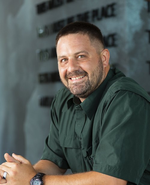 A smiling man in a dark green shirt sits relaxed, his hands clasped together. He appears confident and approachable, sitting in front of a background with the words 'THE PLACE TO BE' in a repeating pattern.