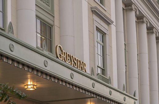 The Greystar logo in elegant gold lettering is mounted on the façade of a building with classic architectural columns, under a softly lit awning, against a dusk sky.