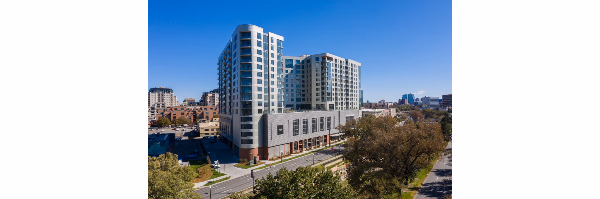 An expansive view of a modern multifamily residential building under clear blue skies. The building has a curved corner with large windows, a rooftop area, and a contrasting lower section with a horizontal siding pattern. It&apos;s situated in an urban area with various other buildings and tree-lined streets in the foreground.