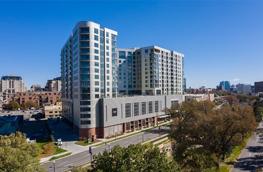 An expansive view of a modern multifamily residential building under clear blue skies. The building has a curved corner with large windows, a rooftop area, and a contrasting lower section with a horizontal siding pattern. It's situated in an urban area with various other buildings and tree-lined streets in the foreground.