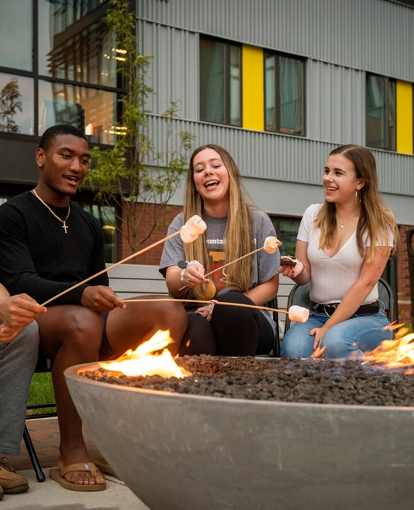Students enjoy a friendly gathering around a fire pit, roasting marshmallows and sharing smiles in a casual outdoor setting.