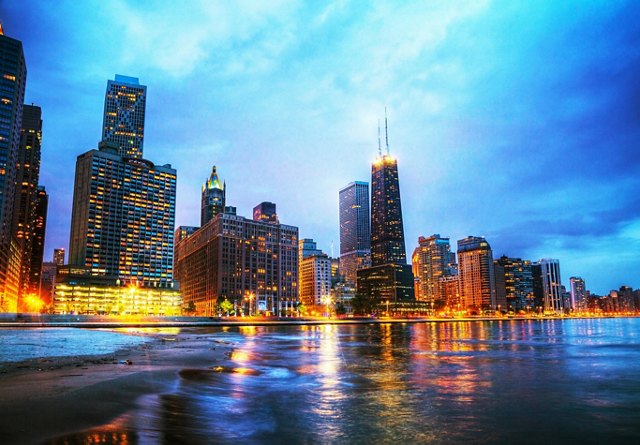 Twilight view of Chicago skyline reflected on the waters of Lake Michigan with lights illuminating the buildings.