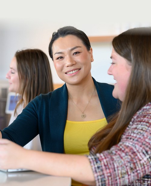 A smiling Asian woman in a business casual attire with two colleagues in conversation in the background.