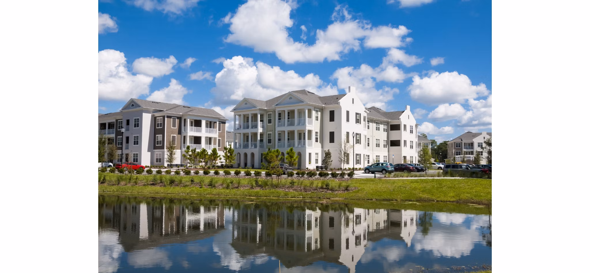 Modern multi-story residential buildings reflected in a serene water body on a sunny day with scattered clouds.
