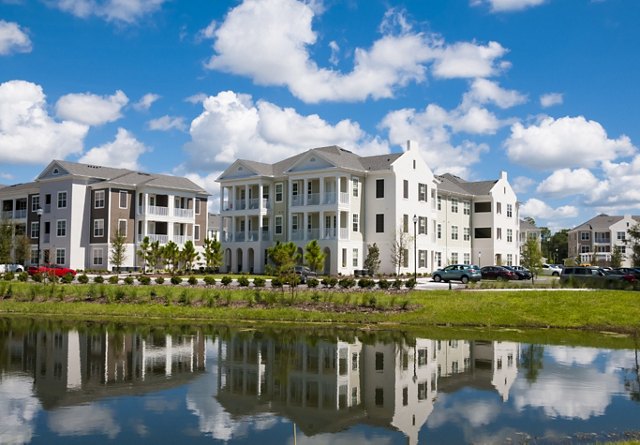 Modern multi-story residential buildings reflected in a serene water body on a sunny day with scattered clouds.