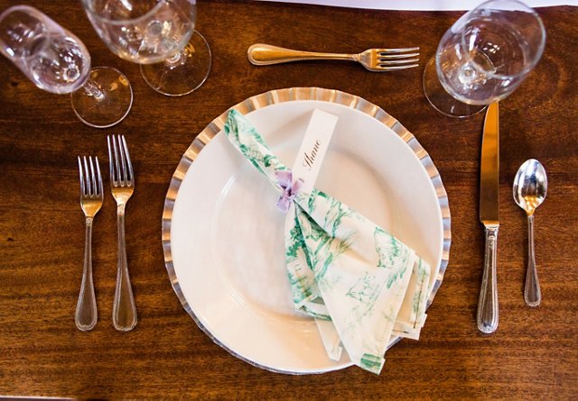 Elegant dining table setting with a folded napkin and name card on a white plate, surrounded by silverware and wine glasses.