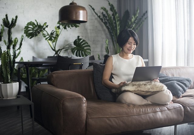 A woman comfortably seated on a leather sofa, working on a laptop in a stylish living room with potted plants and a warm, inviting decor.